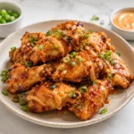 Crispy air fryer chicken wings plated on a ceramic dish with creamy orange sauce drizzle, chopped green onions, and small dipping bowls on a white marble countertop.