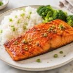 Glazed air fryer salmon fillet served with white rice and steamed broccoli on a ceramic plate, topped with sesame seeds and chopped green onions on a bright marble surface.