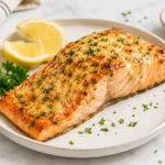 Juicy air fryer salmon fillet topped with garlic herb butter seasoning, served with lemon wedges and parsley on a ceramic plate over a bright marble countertop.