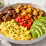 Air fryer breakfast bowl with scrambled eggs, crispy breakfast potatoes, seasoned steak bites, sliced avocado, and diced tomatoes served on a ceramic bowl over a bright marble surface.
