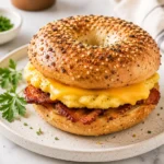 Air fryer breakfast bagel sandwich with fluffy eggs, melted cheddar cheese, and crispy bacon layered inside a seeded bagel on a ceramic plate.
