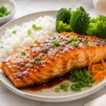 Glazed air fryer salmon fillet served with white rice and steamed broccoli, topped with sesame seeds and green onions on a ceramic plate over a marble countertop.