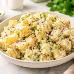 Creamy potato salad served in a ceramic bowl with tender potatoes, herbs, parmesan cheese, red onion, and creamy dressing on a bright marble countertop.