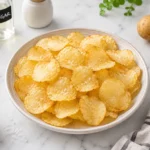 Crispy homemade air fryer potato chips served in a ceramic bowl with flaky salt, golden thin slices, and vinegar seasoning on a bright marble countertop.
