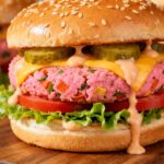 Air fryer pink veggie burger with a vibrant beet based patty, melted cheese, pickles, lettuce, tomato slices, and creamy sauce on a sesame seed bun.