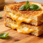 Air fryer grilled cheese sandwich with crispy golden toasted bread and gooey melted cheddar cheese, stacked on a wooden board with fresh basil leaves.