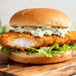 Air fryer fish sandwich with crispy breaded fish fillet, fresh lettuce, and creamy tartar sauce on a soft bun, served with lemon wedges.