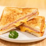 Air fryer ham and cheese sandwich with toasted golden bread, sliced ham, and melted cheddar cheese, served warm on a white plate.