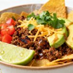Air fryer taco bowl filled with seasoned ground beef, tortilla chips, shredded cheddar cheese, avocado slices, diced tomatoes, fresh cilantro, and lime wedges.