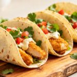 Air fryer chicken tacos with crispy breaded chicken, fresh lettuce, diced tomatoes, cilantro, and creamy sauce in soft tortillas, served with lime wedges.