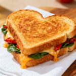 Air fryer heart shaped sandwich with crispy golden toasted bread, melted cheese, fresh spinach, and tomatoes, served as a cute and comforting meal.
