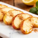 Air fryer apple slices coated with cinnamon and lightly caramelized, served on a platter as a warm and sweet snack with fresh mint garnish.