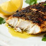 Air fryer blackened fish with a bold spicy seasoning crust, flaky white fish interior, and a drizzle of butter, served with lemon wedges and fresh parsley.