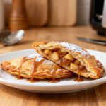 Air fryer fried apple pies with a crispy golden pastry crust, warm cinnamon apple filling, powdered sugar, and caramel drizzle, served as a cozy homemade dessert.