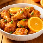 Air fryer frozen orange chicken with crispy breaded chicken pieces coated in a glossy orange sauce, garnished with green onions and fresh orange slices in a serving bowl.