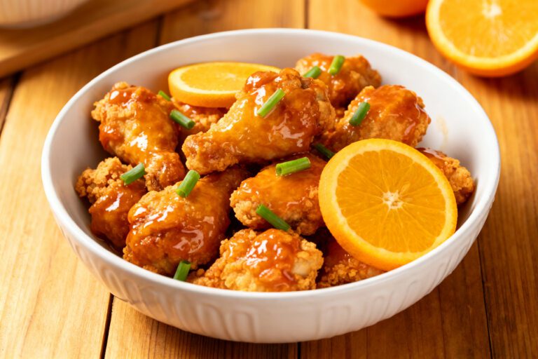 Air fryer frozen orange chicken with crispy breaded chicken pieces coated in a glossy orange sauce, garnished with green onions and fresh orange slices in a serving bowl.