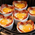 Air fryer heart shaped bacon and egg cups with crispy bacon edges and baked eggs, served in heart shaped ramekins for a fun and festive breakfast.