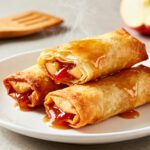 Air fryer apple pie egg rolls with a crispy golden wrapper, warm cinnamon apple filling, and sweet caramel drizzle, served as a cozy homemade dessert.