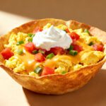 Air fryer egg tortilla bowl with crispy tortilla shell, fluffy scrambled eggs, melted cheese, diced tomatoes, green onions, and a dollop of sour cream, served as a flavorful breakfast.