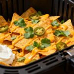 Cheese nachos in the air fryer with crispy tortilla chips, melted cheese sauce, sliced jalapeños, fresh cilantro, and sour cream, served inside an air fryer basket as a quick appetizer.