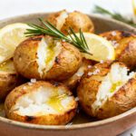 Air fryer Dutch potatoes with crispy golden skins, fluffy cracked centers, flaky sea salt, fresh rosemary, lemon slices, and a drizzle of olive oil, served as a comforting side dish.