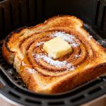 Cinnamon toast in the air fryer with crispy golden bread, swirls of cinnamon sugar, melted butter, powdered sugar, and toasted edges, cooked inside an air fryer basket.