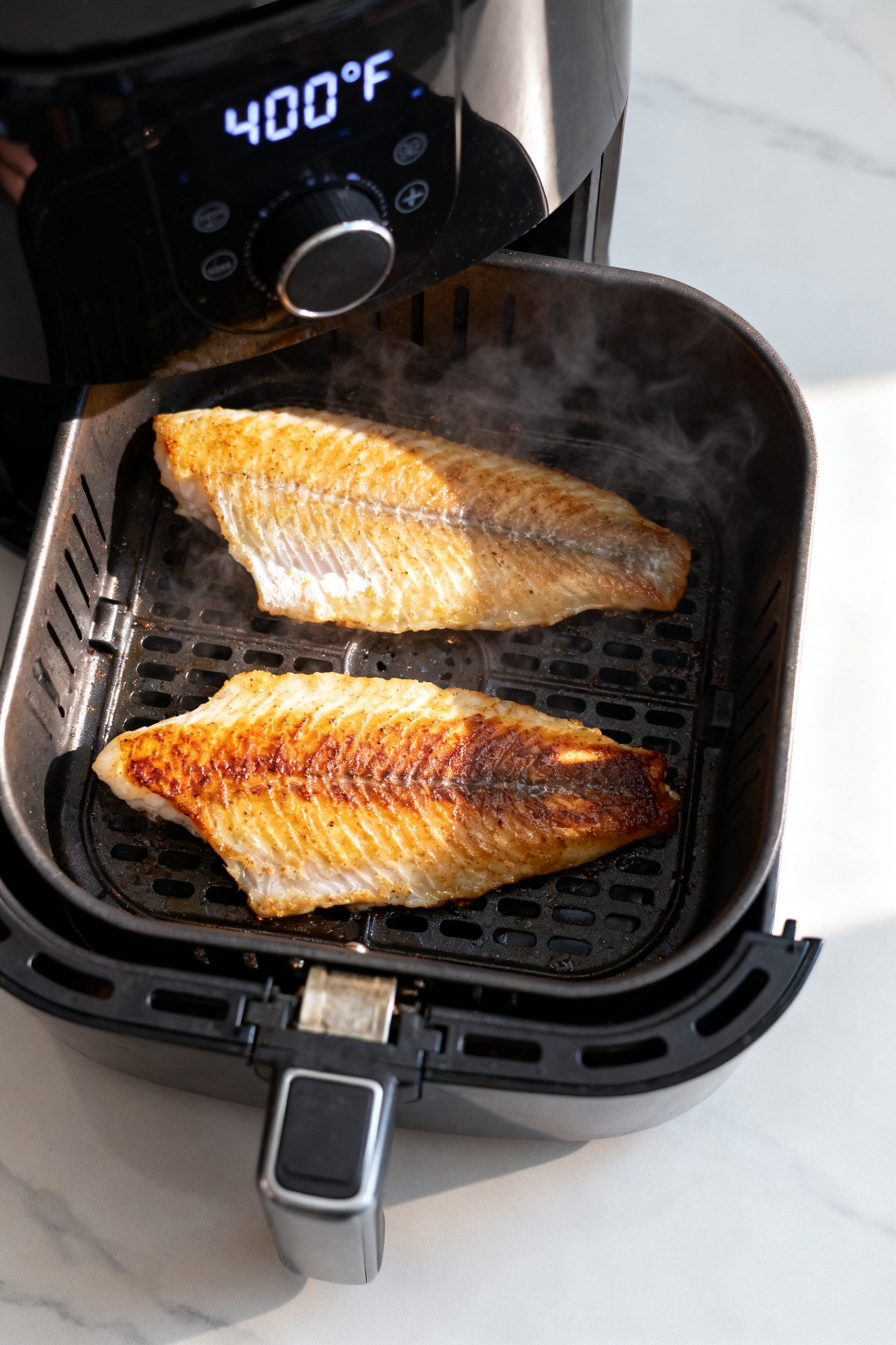 Cooking process: Air fryer basket shot at mid-cook, overhead view of two tilapia fillets arranged in