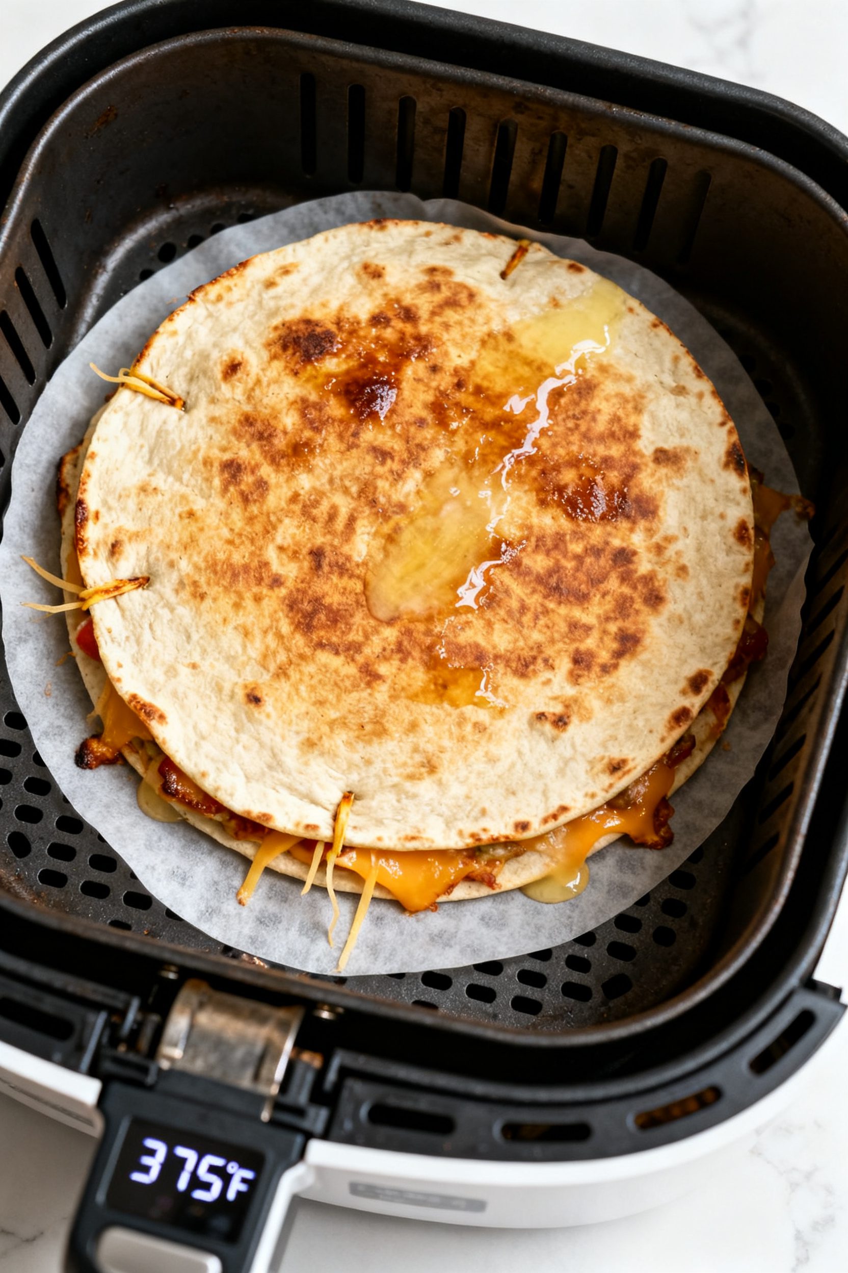 Cooking process: Overhead shot of a full, round quesadilla inside an air fryer basket mid-cook after