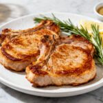 Air fryer keto boneless pork chops with juicy golden browned pork chops, fresh rosemary sprigs, lemon wedges, and a creamy mustard sauce, served as a flavorful low carb dinner.