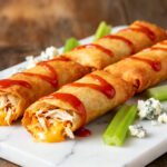 Air fryer buffalo chicken egg rolls with crispy golden wrappers, shredded buffalo chicken, melted cheddar cheese, buffalo sauce drizzle, celery sticks, and blue cheese crumbles, served as a spicy appetizer.