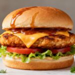 Air fryer chicken burger with a juicy crispy chicken patty, melted cheddar cheese, fresh lettuce, tomato slices, and barbecue sauce on a toasted bun, served as an easy homemade burger.