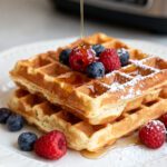 Air fryer waffles with crispy golden edges, fluffy interiors, fresh raspberries, blueberries, powdered sugar, and maple syrup, served as an easy homemade breakfast.