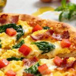 Air fryer breakfast pizza with fluffy scrambled eggs, crispy bacon, fresh spinach, diced tomatoes, melted cheese, and a golden pizza crust, served as a hearty homemade breakfast.