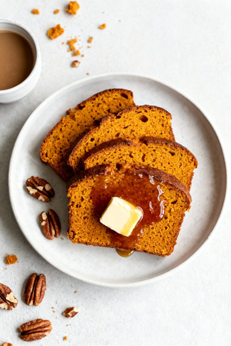 Homemade pumpkin bread with moist golden orange slices, melted butter, maple syrup, pecans, and a warm cup of coffee, served as a cozy fall breakfast or snack.